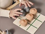 Et barns hænder spiller tic-tac-toe med svampeformede brikker på en træplade med et grønt gitter.