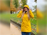 En person i en gul regnfrakke står udenfor på en sti og holder sin hætte, med efterårstræer og blade i baggrunden.