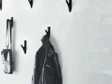En sort læderjakke og en lille sort taske hænger på minimalistiske sorte knager på en lysegrå struktureret væg.
