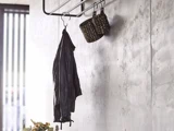 En sort jakke og to flettede kurve hænger på kroge fra et vægstativ i metal mod en lysegrå struktureret væg.