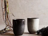 To keramiske kopper, en mørk og en lys, står på en neutral overflade med tørrede kviste og grønt ved siden af på en lys struktureret baggrund.