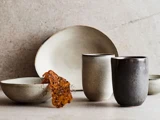 Minimalistisk keramikservice med to tallerkener, to kopper og en dekorativ crisp arrangeret på en neutral stenoverflade.