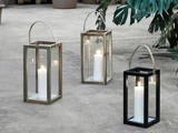 Tre glaslanterner med hvide lys indeni står på en betonflade, omgivet af grønne blade og potteplanter i baggrunden.