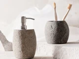 To badeværelsestilbehør med stenstruktur: en sæbedispenser og en kop med to tandbørster af træ, placeret på en hvid struktureret overflade.