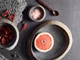 En skive grapefrugt på en stentallerken, med skåle med rosa salt og tørret frugt i nærheden, alt sammen på en mørk, struktureret overflade.
