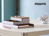 Stablede grå og kork skrivebordsarrangører med kontorartikler på et træskrivebord, ved siden af en indpakket vare og en rød kuglepen. MUUTO-logoet vises i øverste højre hjørne.