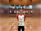 En kvindelig håndboldspiller står på en træbane iført en hvid og rød holdtrøje med sponsorlogoer og nummer 27.