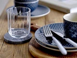 Et klart glas på en bordskåner ved siden af blåmønstrede tallerkener, en kop og gaffel og kniv med hvidt skaft på et træbord.