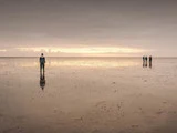 En person står alene på en stor, reflekterende strand ved solnedgang, mens fire andre går sammen i det fjerne under en overskyet himmel.