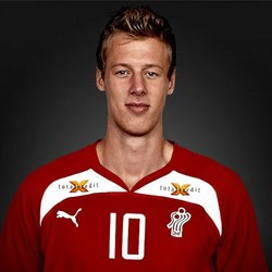 En mand i en rød sportstrøje med nummer 10 og hvide logoer står på en mørk baggrund og kigger ind i kameraet.