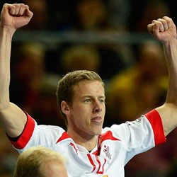 En mandlig atlet i en hvid sportstrøje med røde detaljer løfter begge arme i jubel med et fokuseret udtryk i ansigtet.