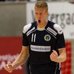 En mandlig håndboldspiller i en mørk trøje jubler med knyttede næver og åben mund efter et spil og viser intense følelser.