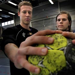 To unge mænd indendørs, den ene i fokus, begge griber en slidt gul og sort håndbold med begge hænder og ser direkte ind i kameraet.