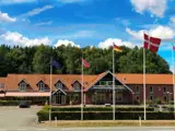 Et hotel med rødt tegltag viser europæiske, norske, tyske og danske flag foran, med træer og blå himmel i baggrunden.