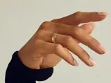 En hånd med lange, velplejede negle bærer en guldring med struktur på langfingeren, set mod en ensfarvet baggrund. Personen er iført en sort ærme.