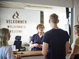 En kvinde står bag en receptionsskranke og taler med tre personer; på et skilt bag hende står der Velkommen på flere sprog.