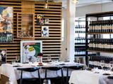 Et moderne restaurantinteriør med hvide duge, dækkede borde, sorte stole, vægkunst og hylder fyldt med vinflasker.