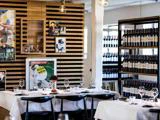 En moderne restaurant med hvide duge, sorte stole, vinflasker på hylder og kunstplakater på en væg af trælameller. Bordene er dækket med glas, tallerkener og servietter.