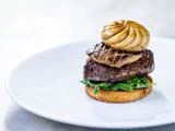 En gourmetret med en ristet rund bund toppet med sauterede grøntsager, en stegt bøf, foie gras og en champignon, arrangeret i en pæn stak på en hvid tallerken.