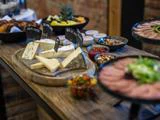 Et træbord med forskellige slags ost, mærket med skilte, skåle med nødder, frugt, dip og tallerkener med skiveskåret kød og salat er opstillet som en buffet.