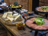 Et træbord med forskellige oste, kød, saucer, nødder og en skål med friske grøntsager er opstillet som en buffet.