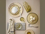 Flat-lay af en minimalistisk borddækning med glasskåle, en kande, en kop, hvidløgsløg, nudler, en gaffel, en ske, en serviet og en træbakke på en beige baggrund.