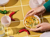 Hænderne holder en skål nudelsuppe på et gult flisebord med røde og grønne chilipeber, kopper, tallerkener og en vandflaske.