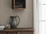 En mørk keramikkande, et glas med en lys drink og bøger står på et træbord under en væghylde med foldet sengetøj ved siden af et gennemsigtigt gardin, som sollyset skinner igennem.