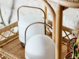 To moderne hvide lanterner med buede metalhåndtag står på en flettet rattanbakke, og en del af en flettet kurv og møbler er synlige i nærheden.