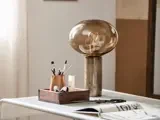 En moderne bordlampe med en rund, ravfarvet glasskærm står på et skrivebord ved siden af en trækasse med pensler og kunstartikler.
