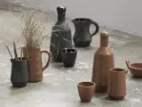 Forskellige keramiske kar i brune og sorte toner, bl.a. kopper, kander, flasker og en kande med tørret græs, arrangeret på et struktureret betongulv.