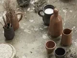 Forskellige keramiske kopper, kander og en vase med tørrede kviste arrangeret på en forvitret betonoverflade. Nogle kopper indeholder væske, andre indeholder pensler eller er tomme.