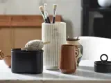 På et marmorbord står en riflet hvid vase med pensler, en sort beholder med sejlgarn, et brunt krus, en mønstret kop med blyanter og en rund sort beholder med sløjfelåg.