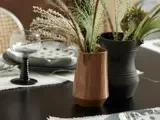 En brun keramikvase med tørret græs og en sort kande med grønt står på et mørkt bord dækket med tallerkener, glas og servietter.