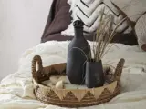 En flettet bakke med en mørk flaske, en vase med tørret græs og en lille dekorativ genstand er placeret på en seng med hvidt og brunt sengetøj.