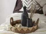 En flettet bakke med en mørk flaske, en vase med tørret græs og en lille dekorativ genstand er placeret på en seng med hvidt og brunt sengetøj.