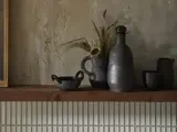 På en træhylde står mørke keramikkar og en kande med tørret græs mod en struktureret beige væg og et hvidt flisebelagt backsplash.