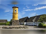 Et gult tårn med 1857 på står på en stensokkel, omgivet af traditionelle stråtækte huse under en blå himmel.