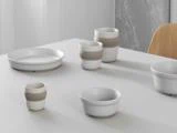 En minimalistisk borddækning med hvide keramiske fade og kopper, nogle med beige bånd, arrangeret på et lyst bord i en moderne indretning.