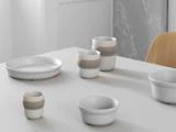 Et minimalistisk hvidt bord med keramiske kopper, skåle og en tallerken, alle med lyse beige bånd, ved siden af en lys træstol.