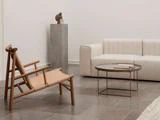Minimalistisk stue med en beige sofa, en træstol, et rundt sofabord med en bog, klinkegulv og en høj grå piedestal med en lille abstrakt skulptur.