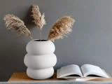 En moderne hvid vase med tre stængler af pampasgræs står på en træplade ved siden af en åben bog på en grå væg.