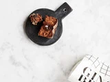 Et stykke mørk kage og et lille stykke af den hviler på et rundt, sort serveringsbræt på en hvid marmoroverflade. En del af en hvid emballage med GIN-tekst er synlig i hjørnet.