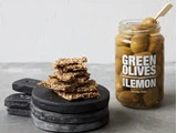 En stak kiks på sorte plader ved siden af et åbent glas med grønne oliven med citron og en lille trægaffel inde i glasset.