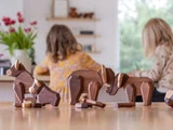 Dyrefigurer af træ på et bord med to børn i baggrunden, der tegner eller skriver, og en vase med blomster i nærheden i et lyst rum.