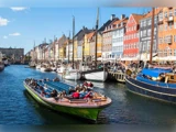 En båd med turister sejler forbi farverige bygninger og fortøjede sejlbåde langs den livlige havnefront i Nyhavn i København på en solrig dag.