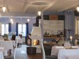 Et hyggeligt restaurantinteriør med hvide duge, en tændt pejs, hylder med glas og flasker og blød pendelbelysning. Blå gardiner indrammer vinduerne i baggrunden.