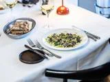 Et restaurantbord dækket med vinglas, bestik, en tallerken salat med grønt og brød på en hvid dug.