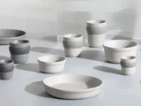 En samling af moderne, minimalistiske keramiske skåle og kopper i hvide og grå nuancer, arrangeret på en lys overflade med en blød baggrund.