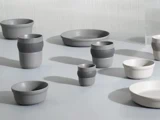 En samling af moderne, minimalistiske keramikskåle og kopper i grå og hvide nuancer arrangeret på en lys overflade.